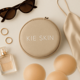 Kie Skin travel pouch flat lay with nipple covers, sunglasses, gold rings, perfume and a satin slip